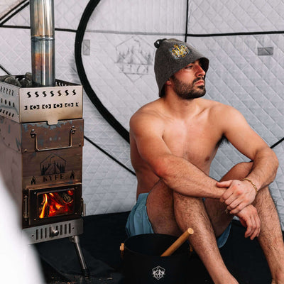 Man relaxing inside arctic gray KYFE portable sauna tent wearing sauna hat beside wood-burning stove with open fire – Finnish wood-fired sauna experience